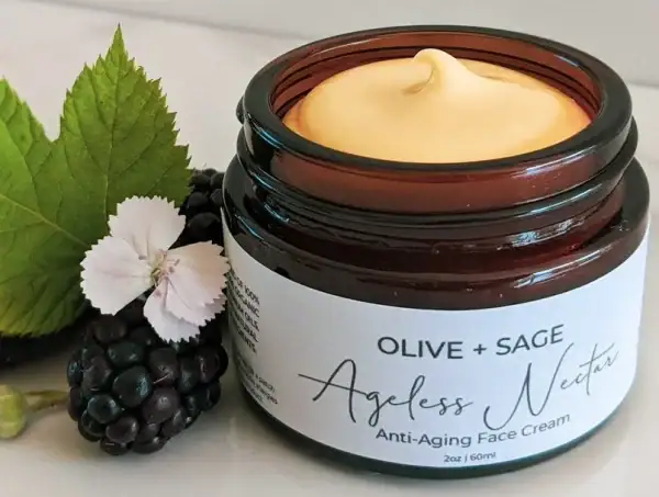 Organic anti aging face cream in glass jar with lid off, showcasing its smooth texture next to fresh blackberries and flowers.