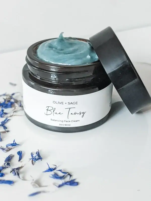 Jar of Organic Blue Tansy Moisturizer surrounded by blue tansy flowers, ideal for calming sensitive skin.