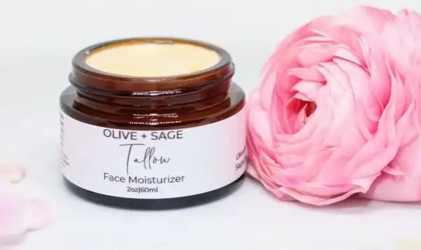 Whipped tallow cream in a glass jar with lid off, accented by a pink rose, showcasing natural ingredients.